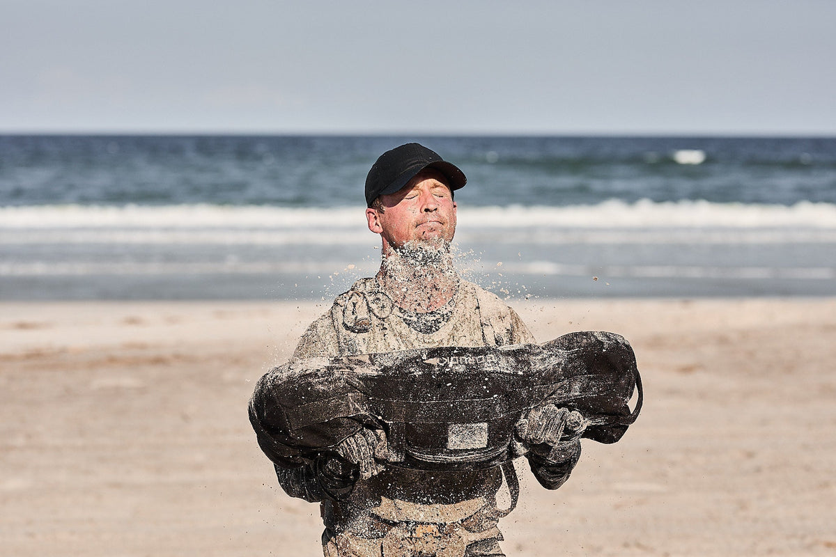 Man lifting GORUCK sandbag on sandy beach, wearing cap and rucking gear near ocean