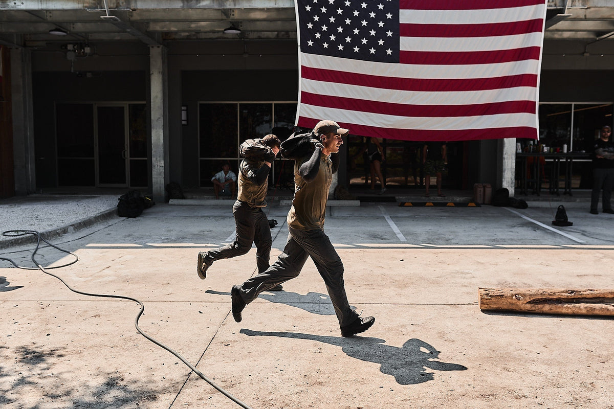 Two people rucking outdoors with weighted bags, American flag, GORUCK training event