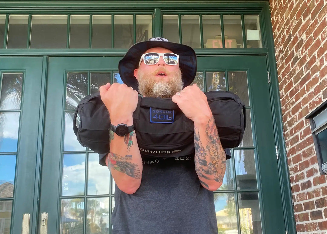 Bearded man with tattoos lifting GORUCK sandbag outdoors, wearing sunglasses and bucket hat