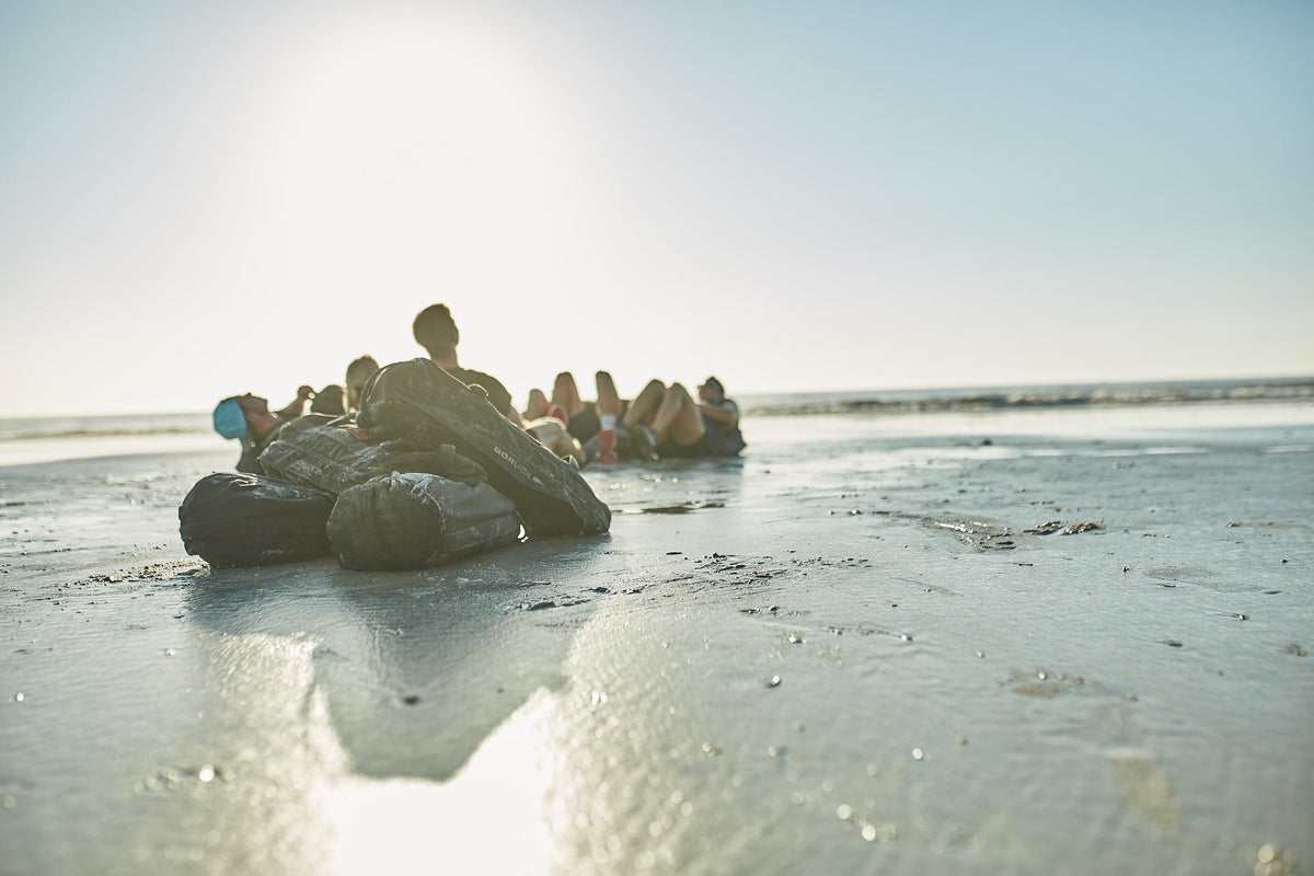 GORUCK sandbags on wet beach with a group training in the background at sunrise