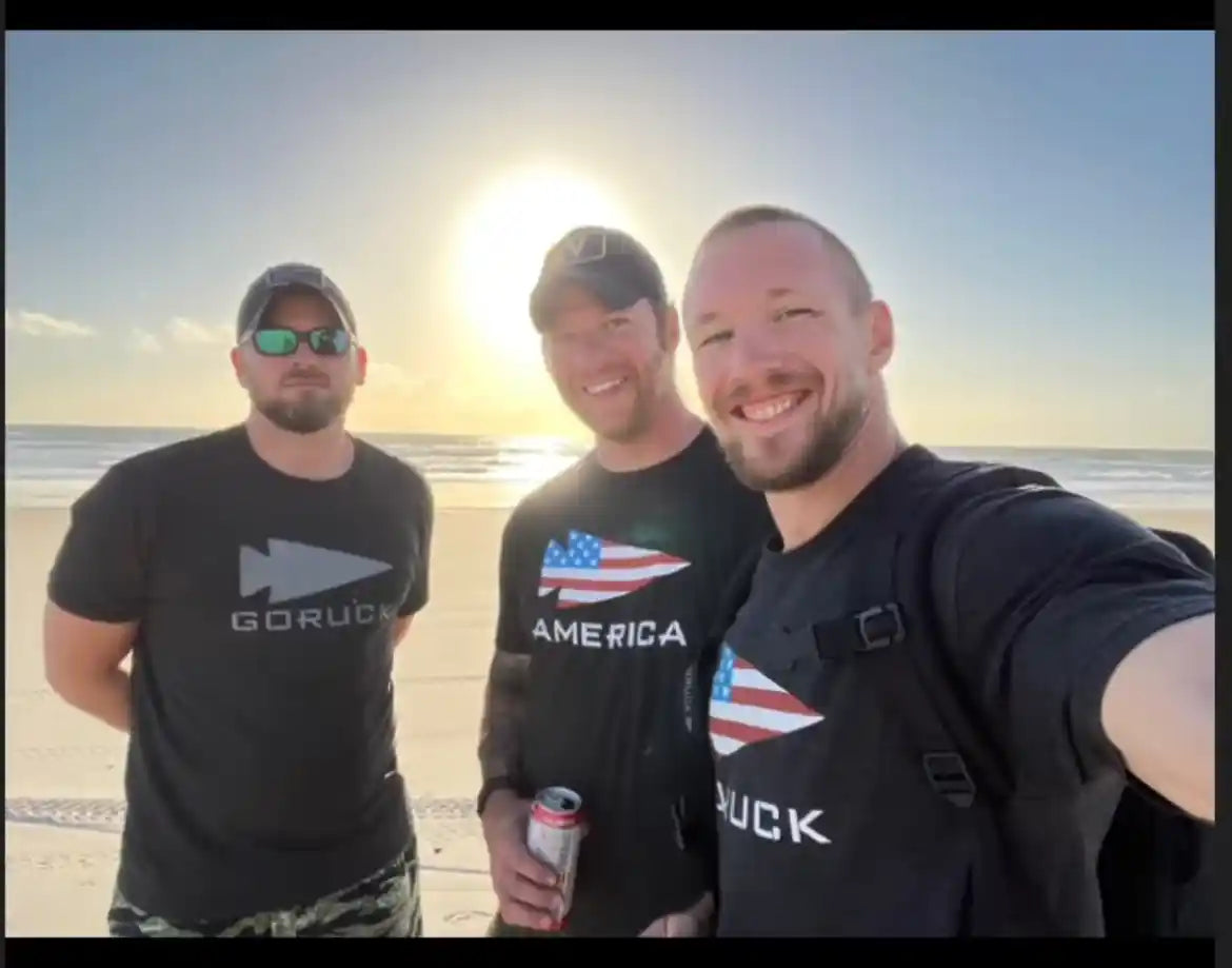 Three men wearing GORUCK shirts smiling on a sunny beach, ocean in background.
