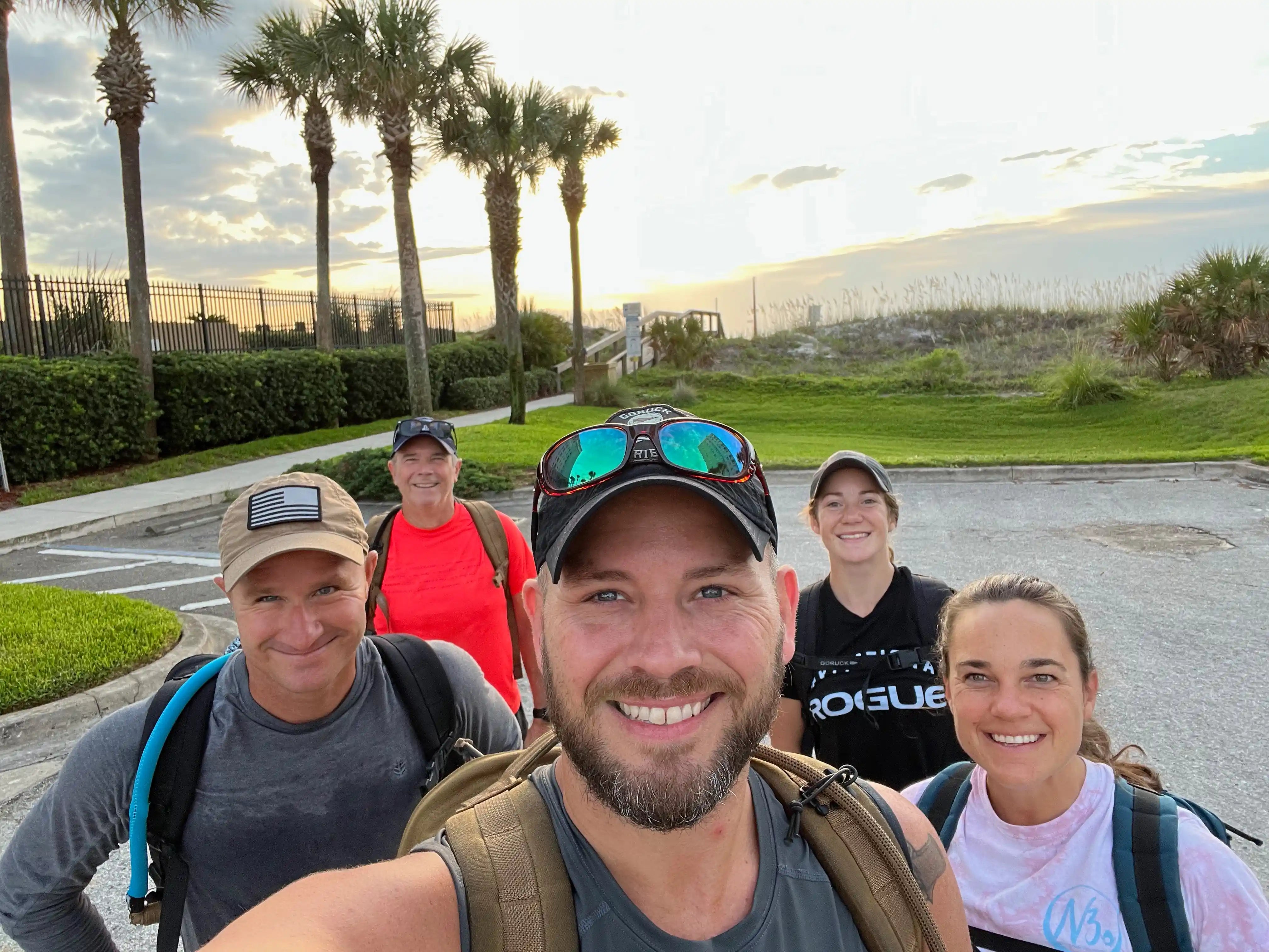 Group of smiling GORUCK ruckers with backpacks outdoors near palm trees at sunrise