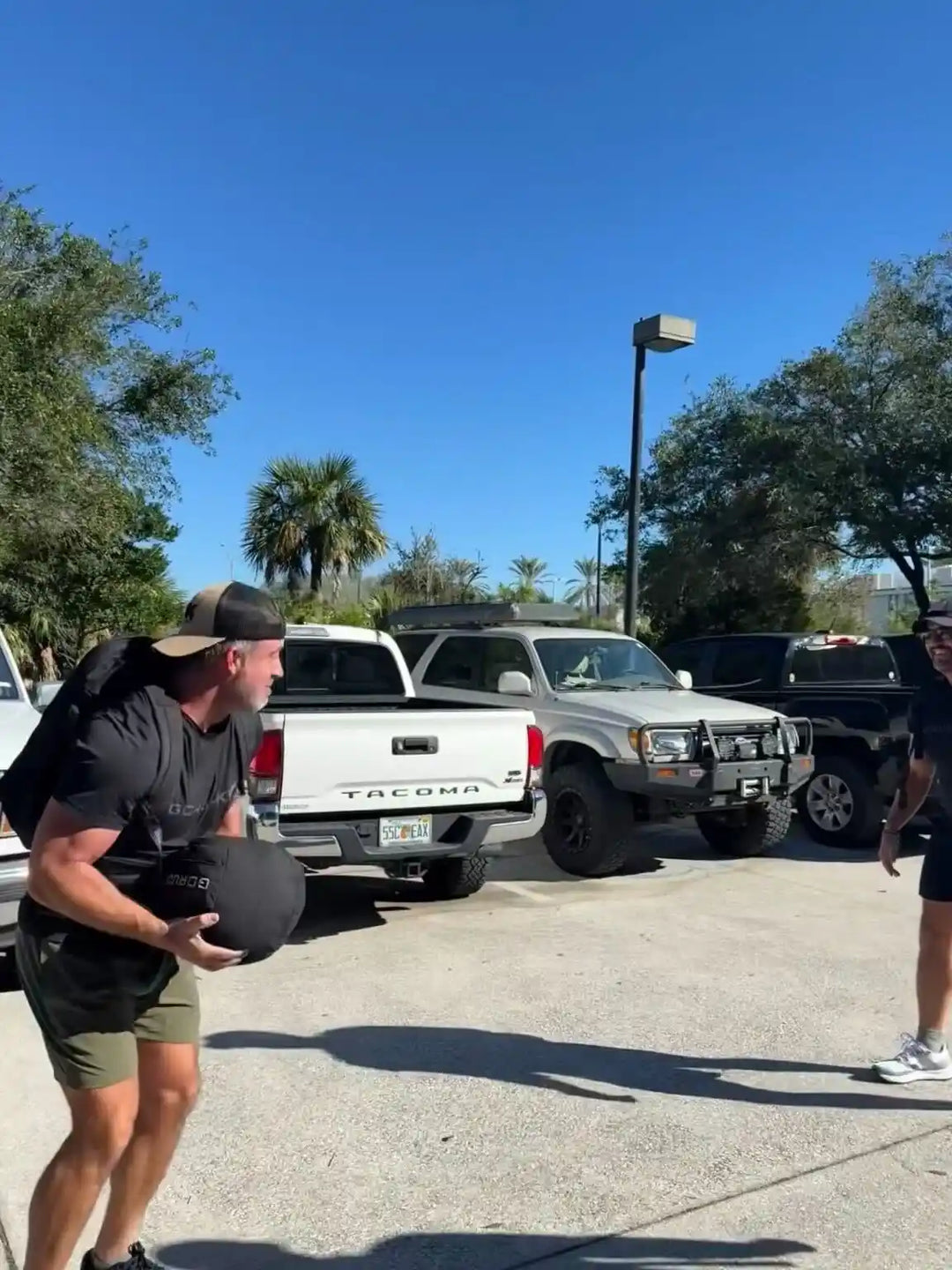 Man training outdoors with a GORUCK sandbag in a parking lot, wearing athletic gear