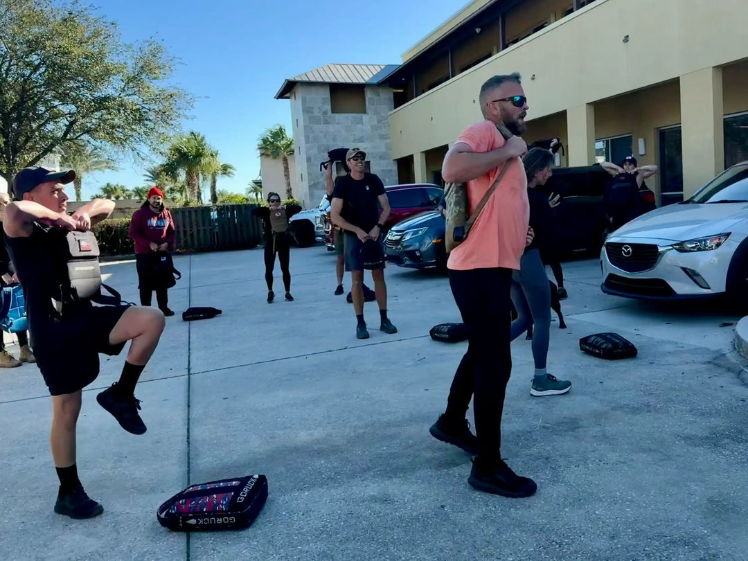 Group outdoor rucking workout with GORUCK gear on sunny day near clubhouse and parked cars
