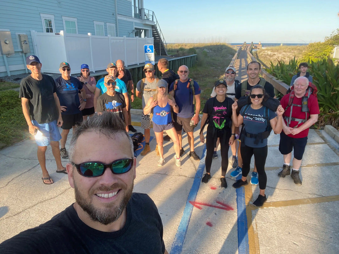 Group of people with rucksacks smiling outdoors near a boardwalk, GORUCK rucking event