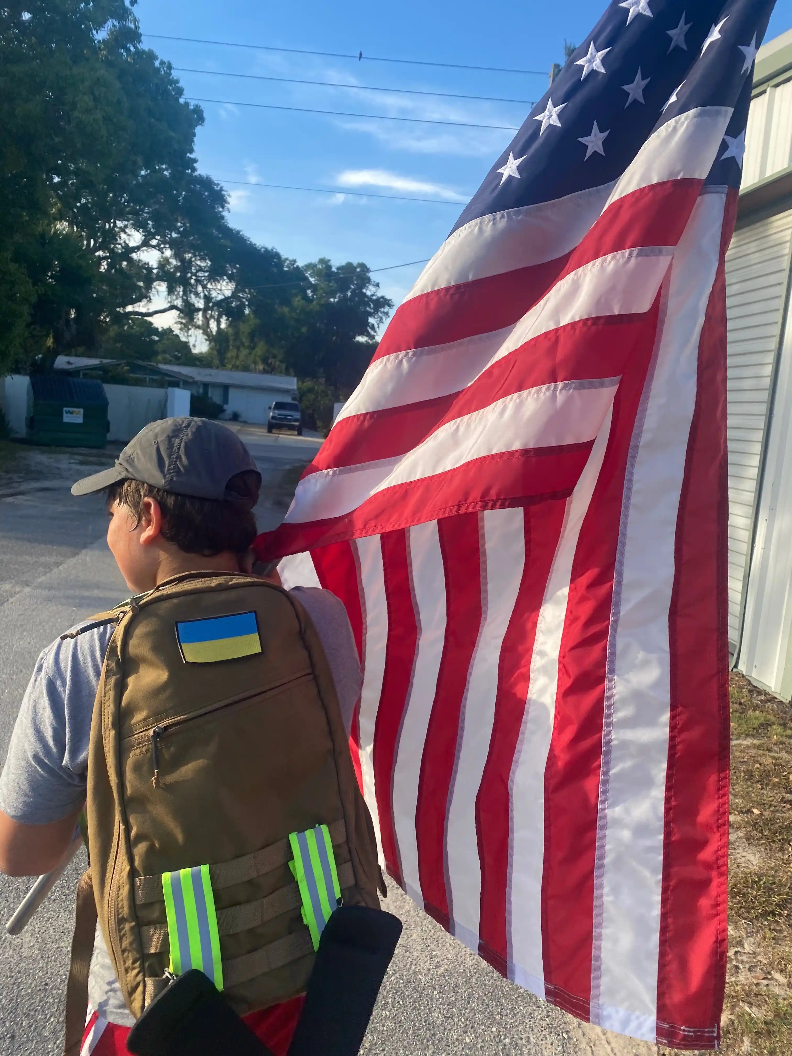 Person rucking with GORUCK backpack and large American flag outdoors on sunny day