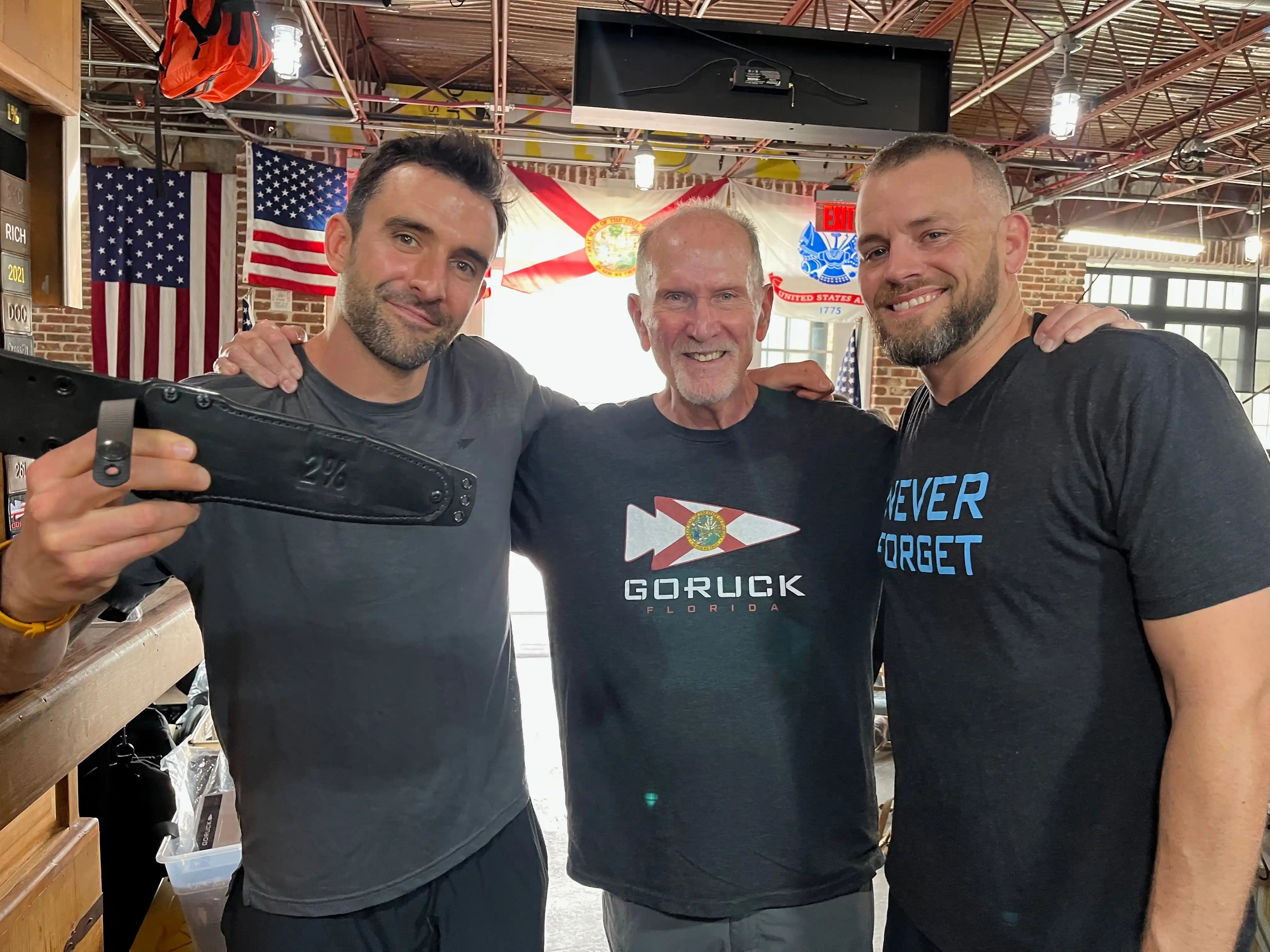Three men at a GORUCK event indoors, with American and Florida flags in the background.