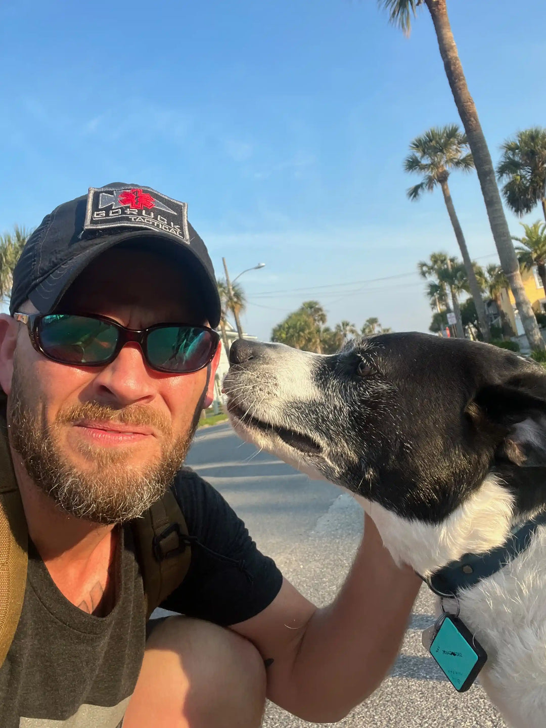 Man wearing GORUCK cap with dog outdoors, sunny day, palm trees in background