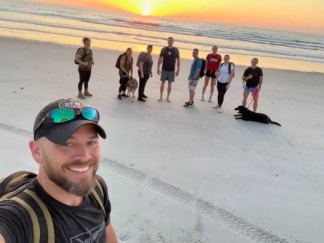 Group of people with backpacks and dogs rucking on the beach at sunset, GORUCK community event