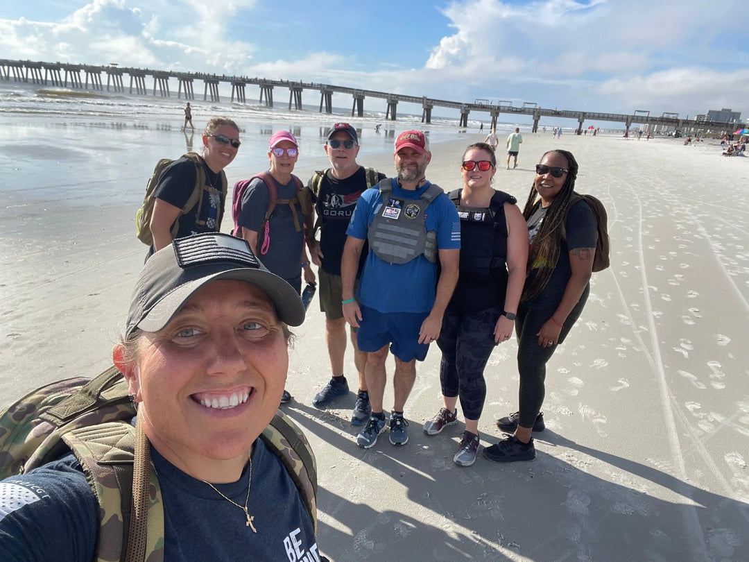 Group of people rucking with GORUCK gear on a sunny beach near a pier.