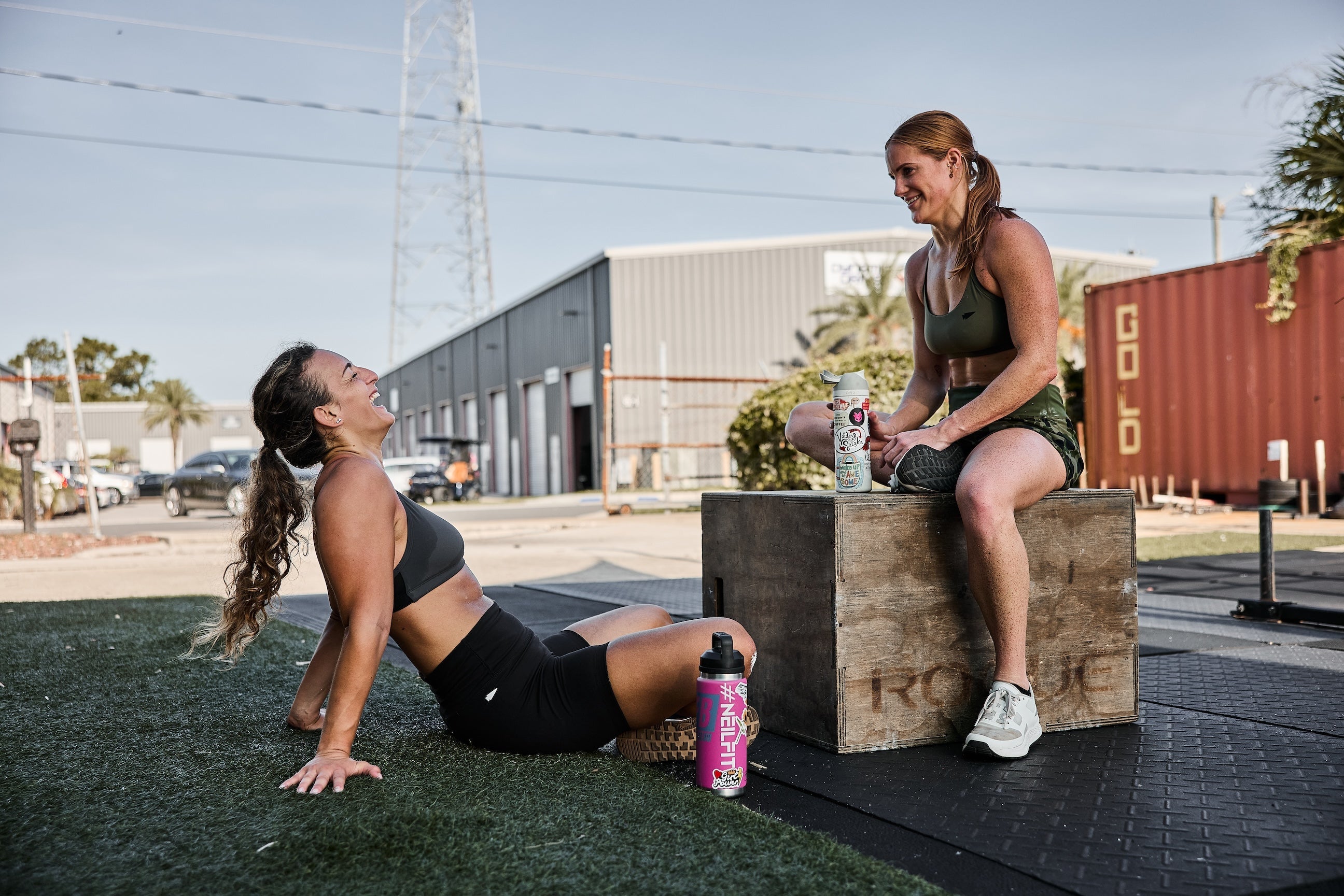 Two women in athletic gear relaxing and laughing after outdoor GORUCK workout by gym equipment