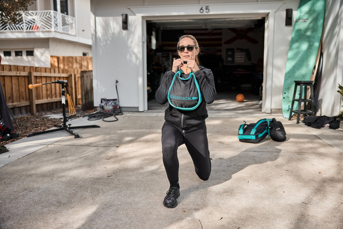Woman performing lunge with GORUCK sandbag outdoors in driveway, wearing black athletic gear