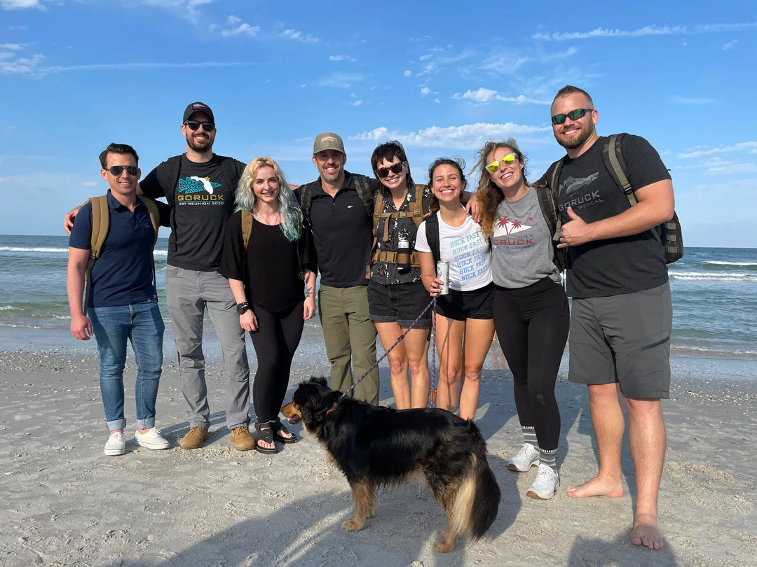 GORUCK team posing with backpacks and a dog on a sunny beach