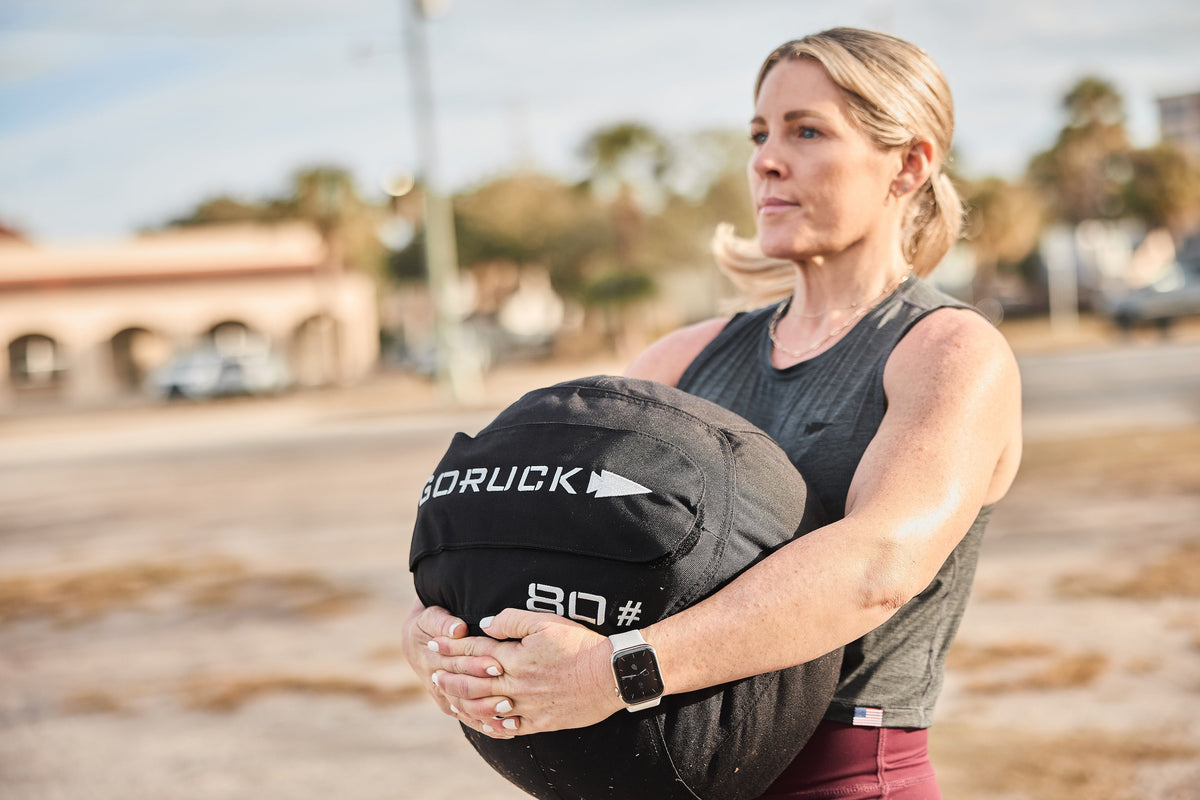 Woman outdoors lifting GORUCK 80 lb sandbag, focusing on strength training with rucking gear.