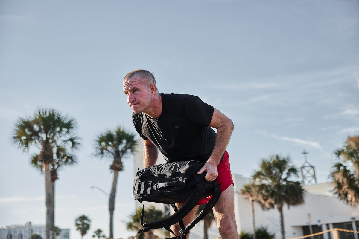 Man outdoors in athletic wear lifting GORUCK backpack, palm trees and sky in background