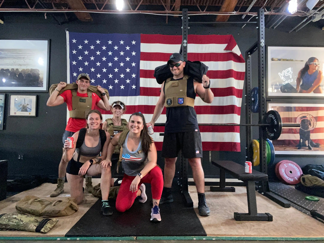 Group wearing GORUCK gear posing in gym with American flag, rucking equipment visible