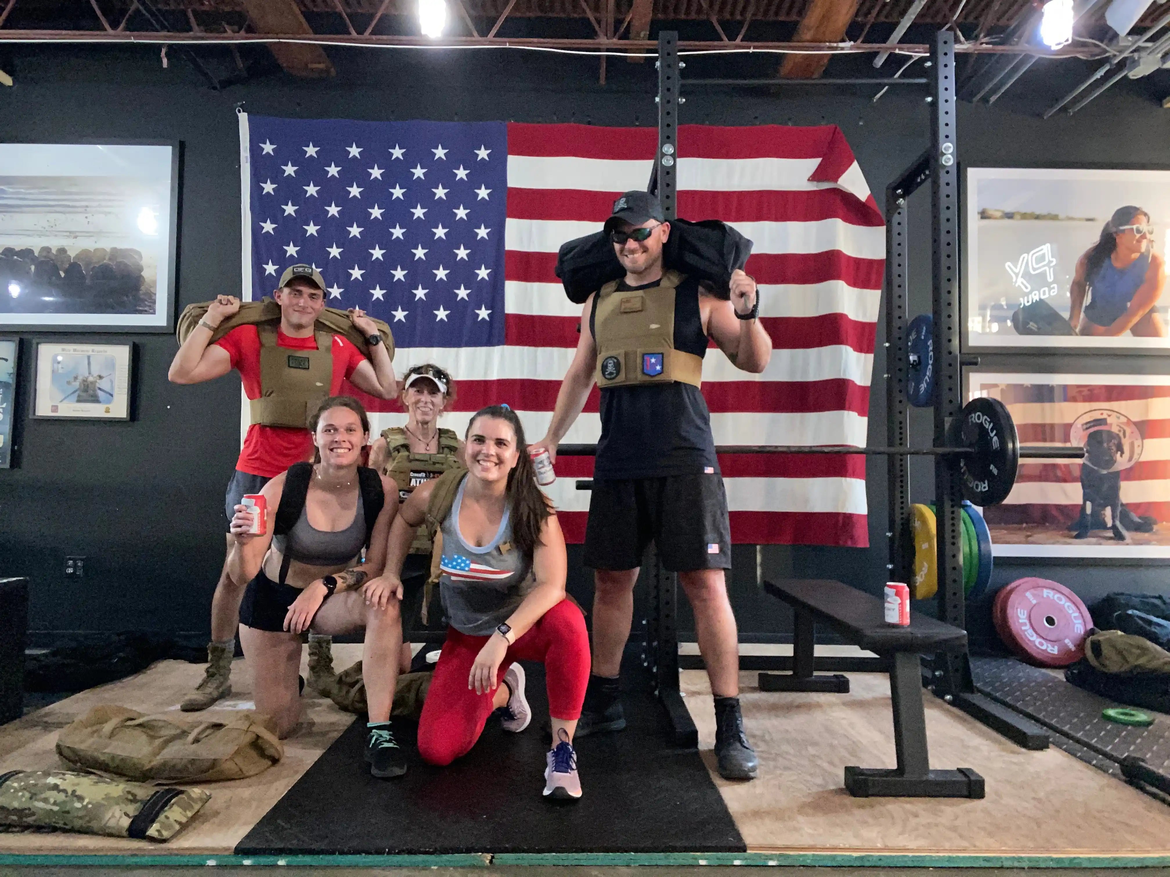 Group wearing GORUCK gear posing in gym with American flag, rucking equipment visible