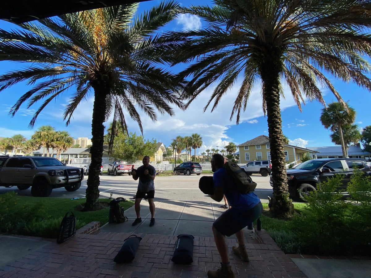 Two people rucking and training with weighted gear between palm trees outside, GORUCK style