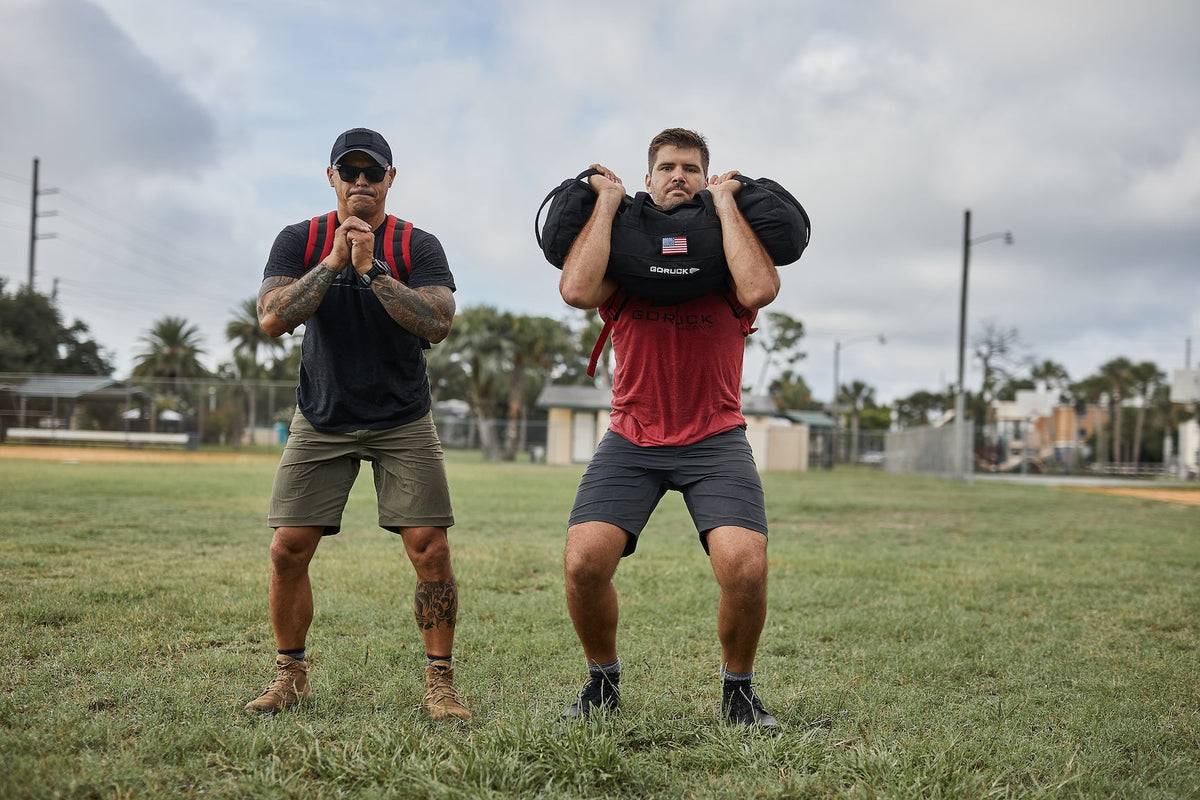 Two men performing outdoor rucking workout with GORUCK sandbags on a grassy field