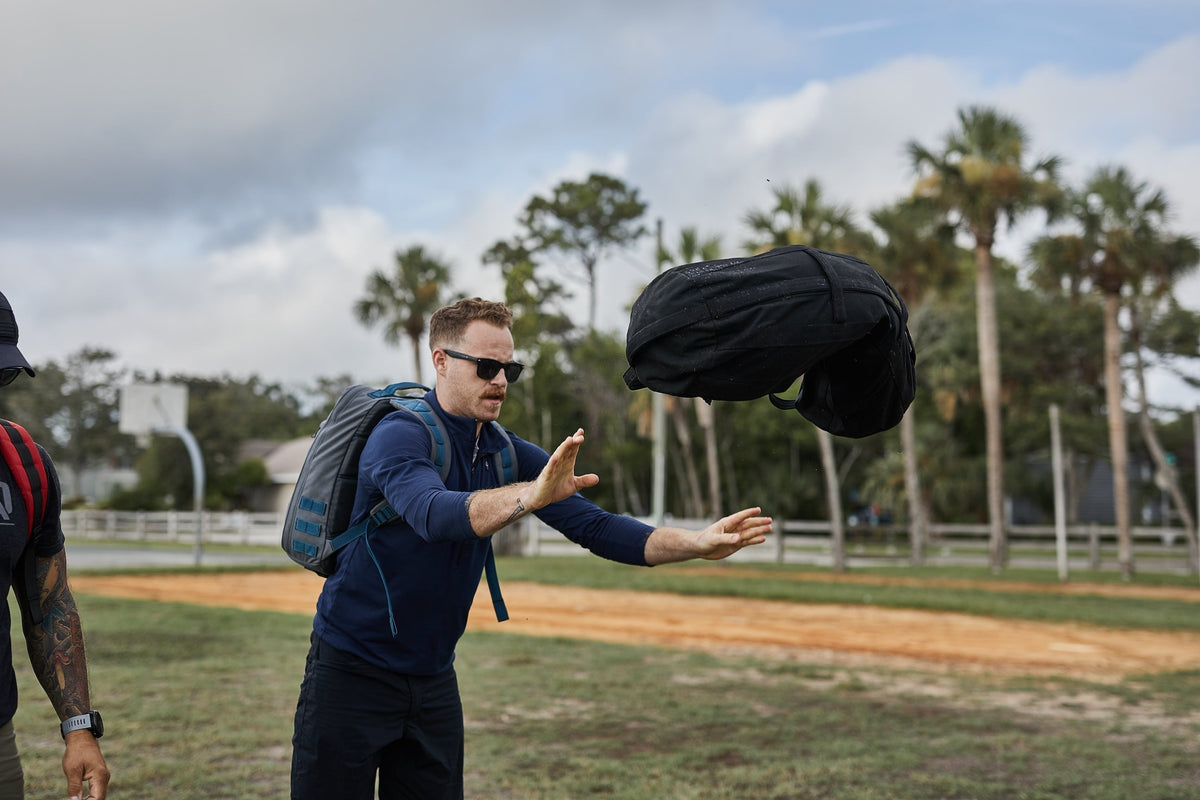 Man tossing GORUCK sandbag outdoors during rucking fitness training with palm trees in background