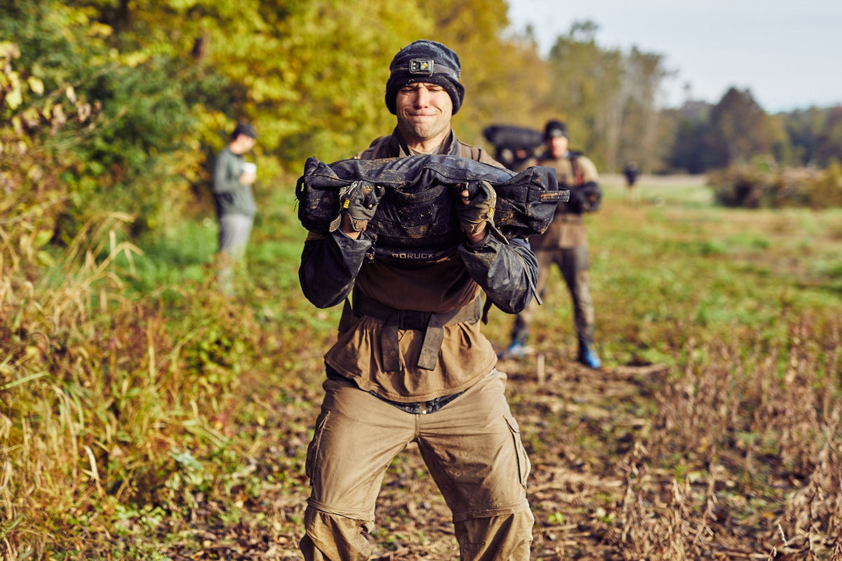Man lifting GORUCK training sandbag outdoors, rucking gear in muddy field