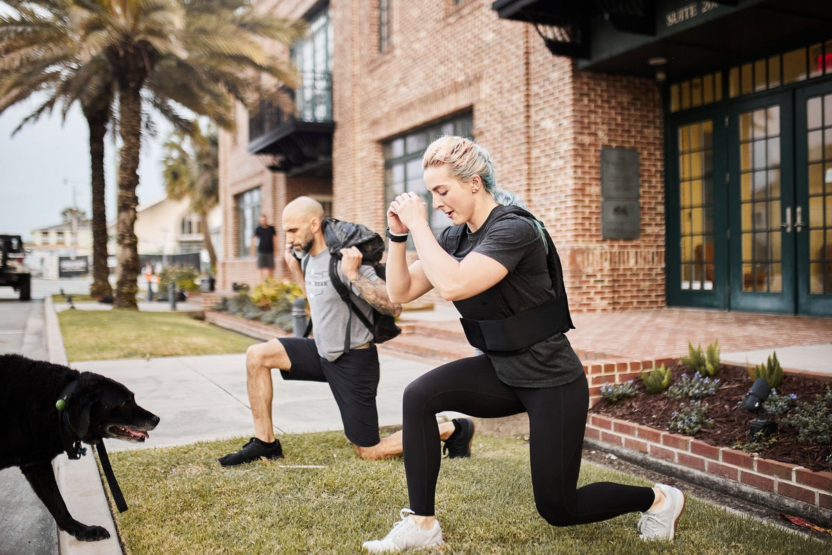 Two people wearing GORUCK weighted vests doing outdoor lunges on grass near a brick building