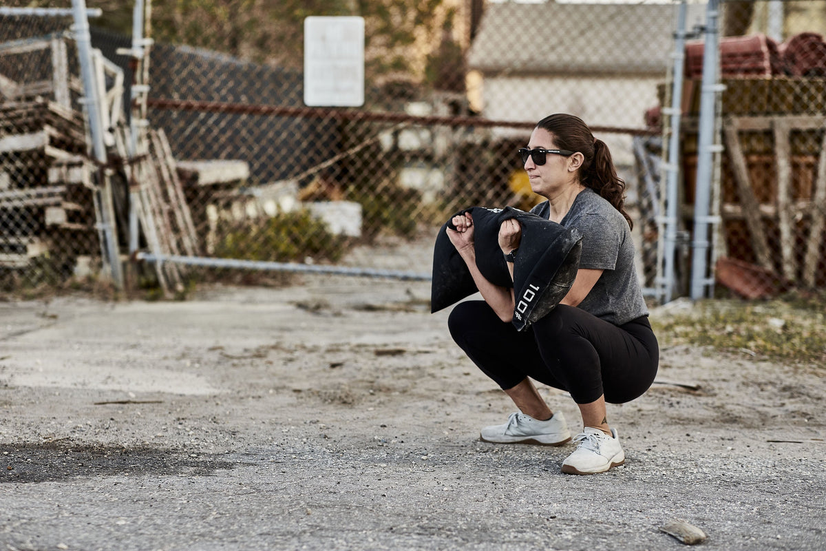 Woman performing ruck squat outdoors using GORUCK sandbag, fitness training in urban yard