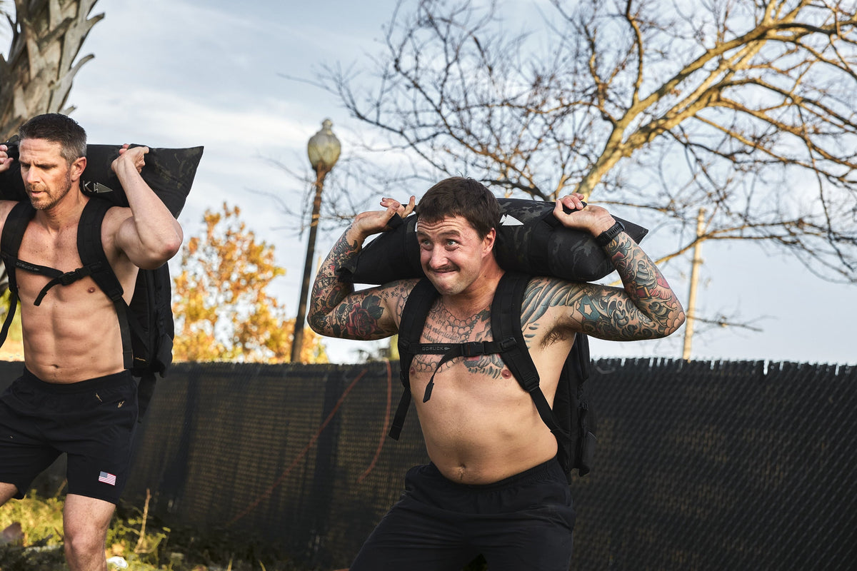 Two men rucking outdoors with GORUCK gear, shirtless, carrying weighted sandbags.