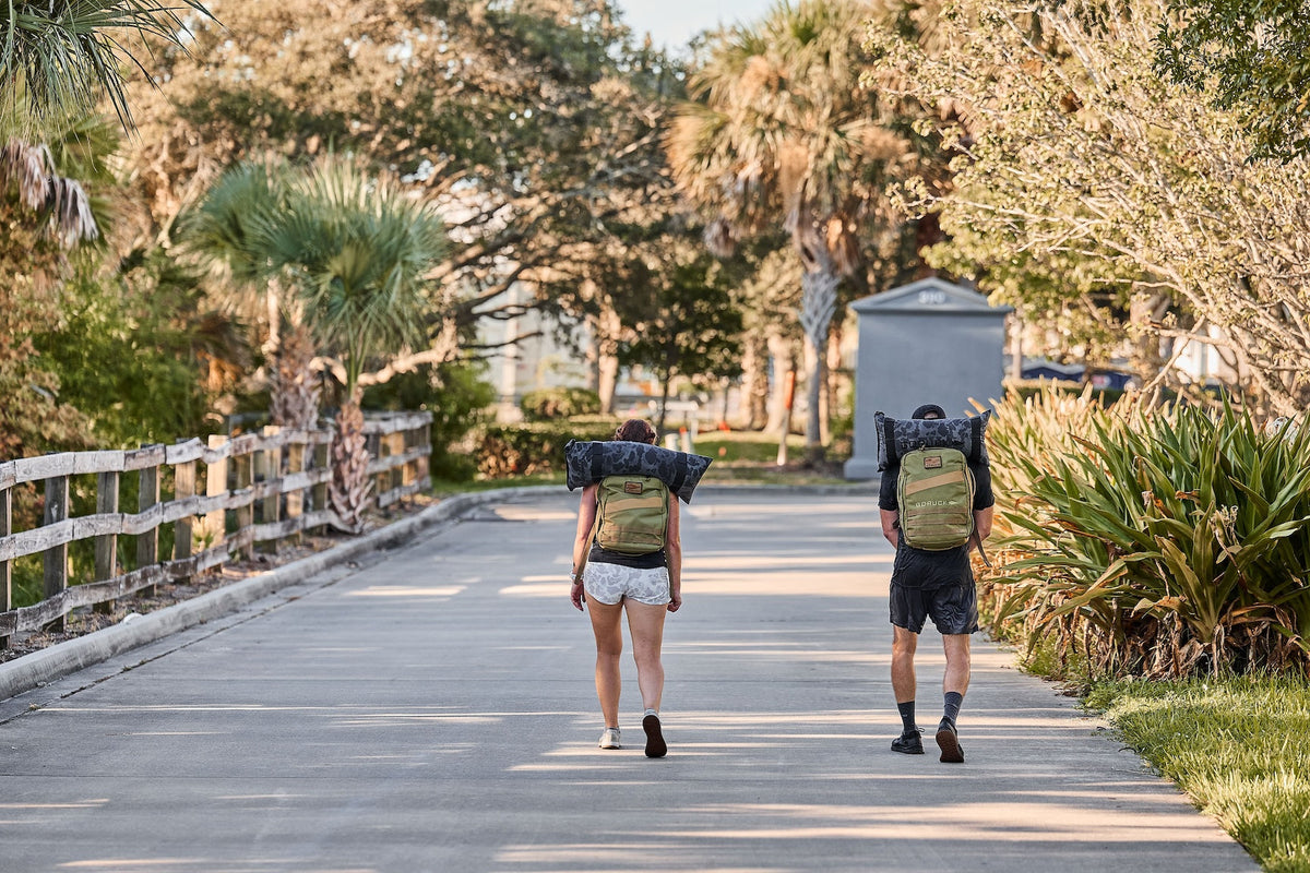 Two people rucking with GORUCK backpacks outdoors on a sunny path lined with palm trees.