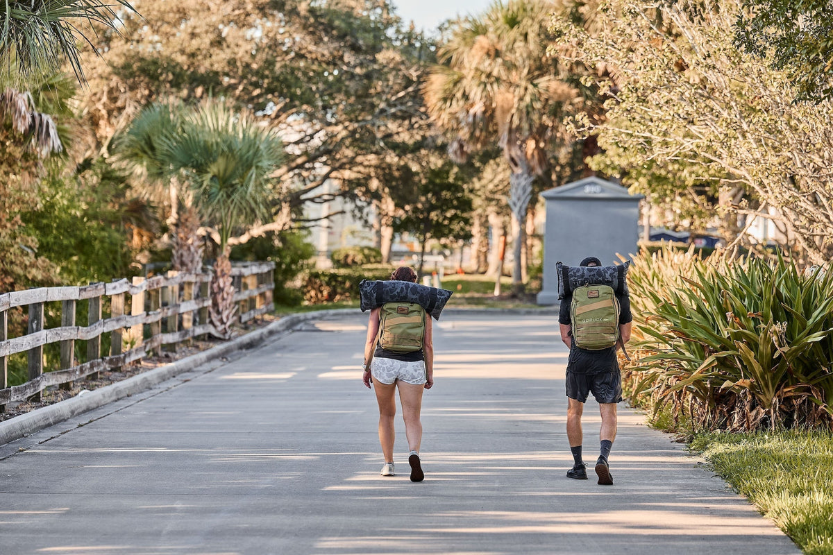 Two people rucking with GORUCK backpacks on a sunlit outdoor path surrounded by greenery.