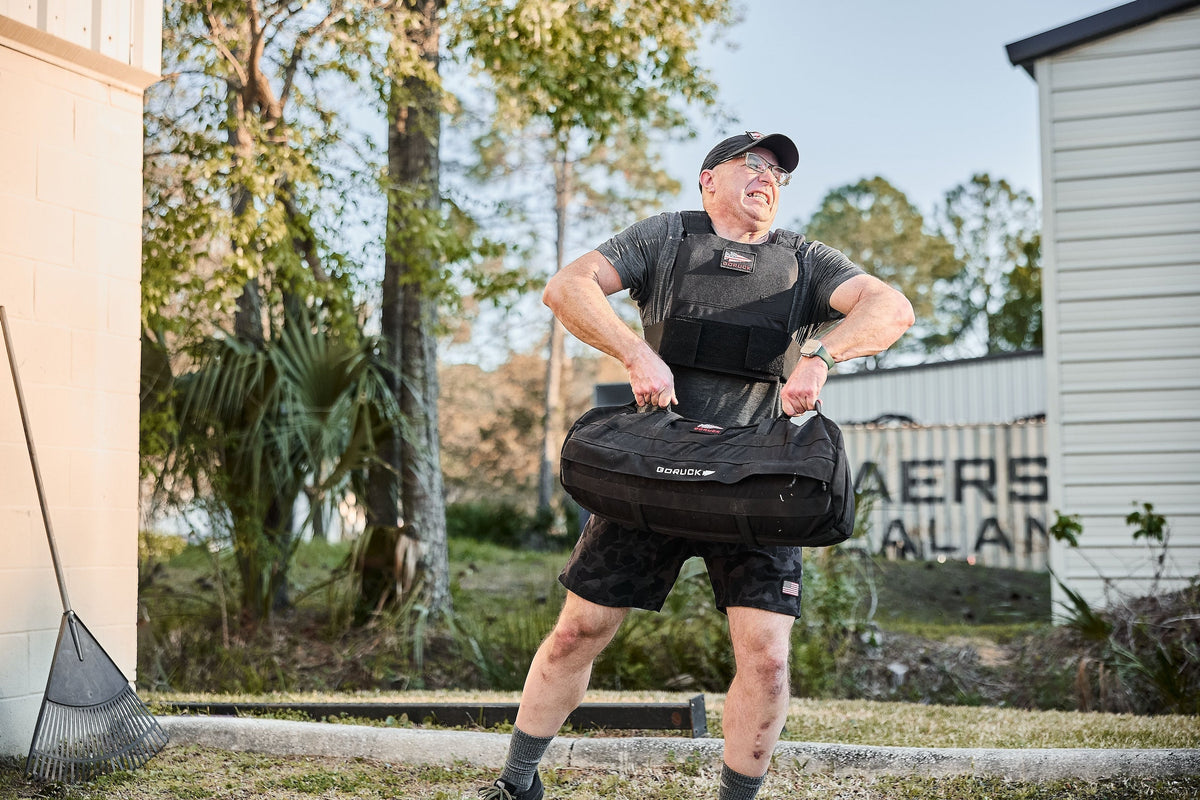Man lifting GORUCK sandbag outdoors, wearing weighted vest, GORUCK rucking gear