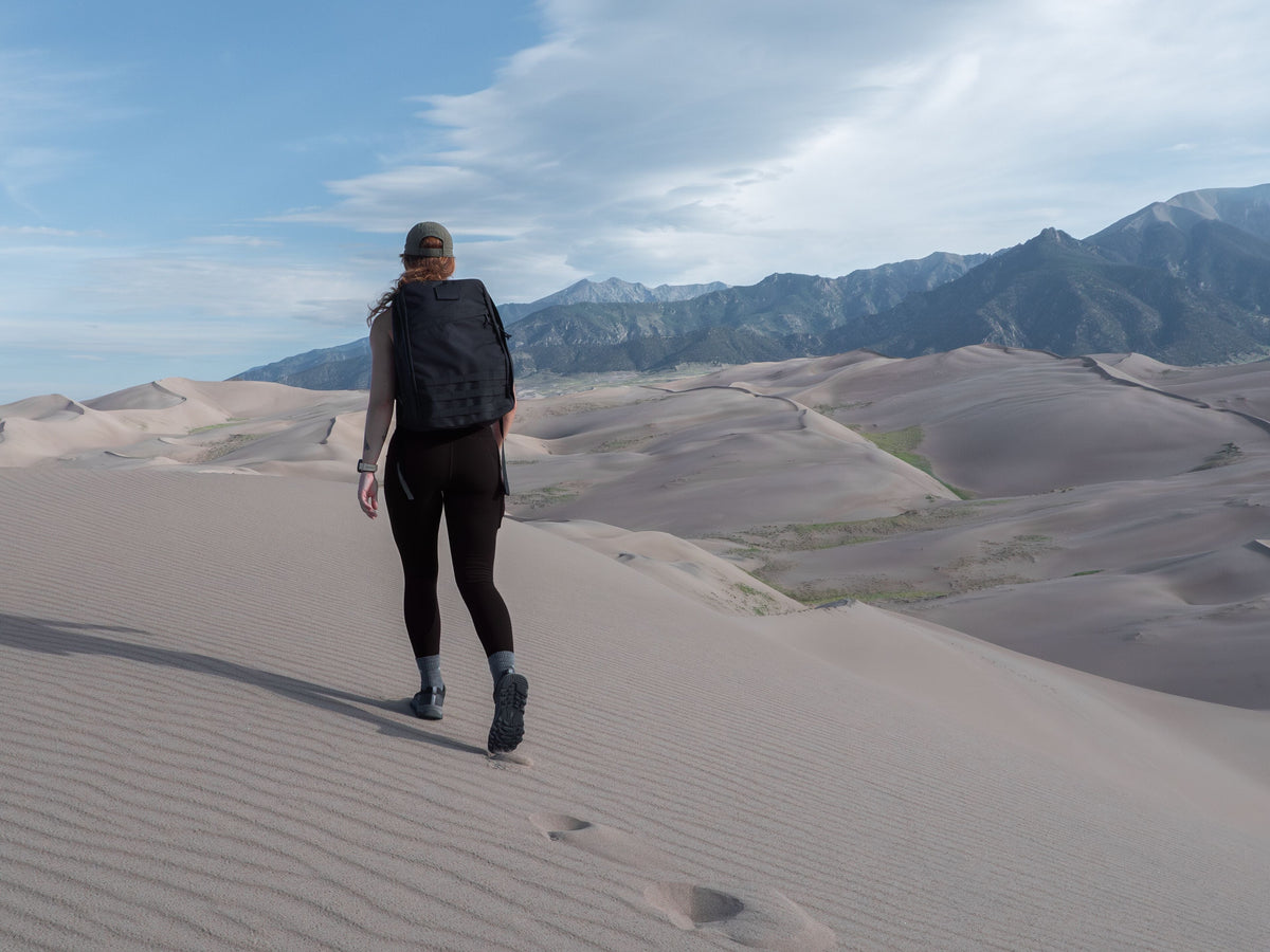 Woman rucking with GORUCK backpack on sand dunes, mountains in background under blue sky