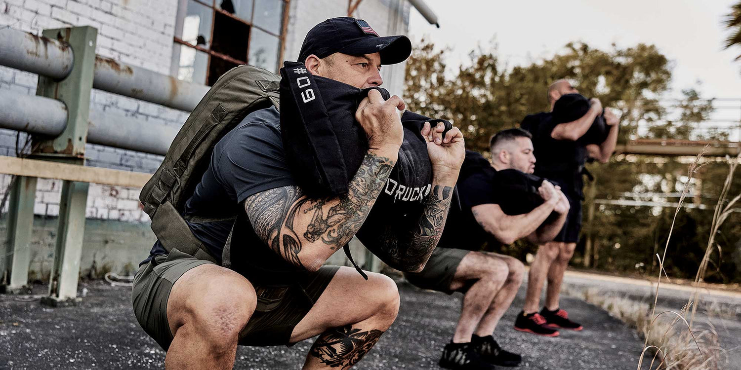 Three men outdoors doing squats, holding sandbags at their chests, next to an industrial building.