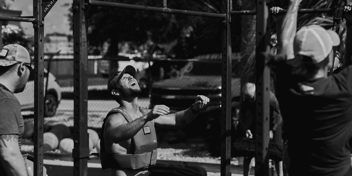Man lifting weights at an outdoor gym, wearing a cap and vest, surrounded by others. Black and white image.