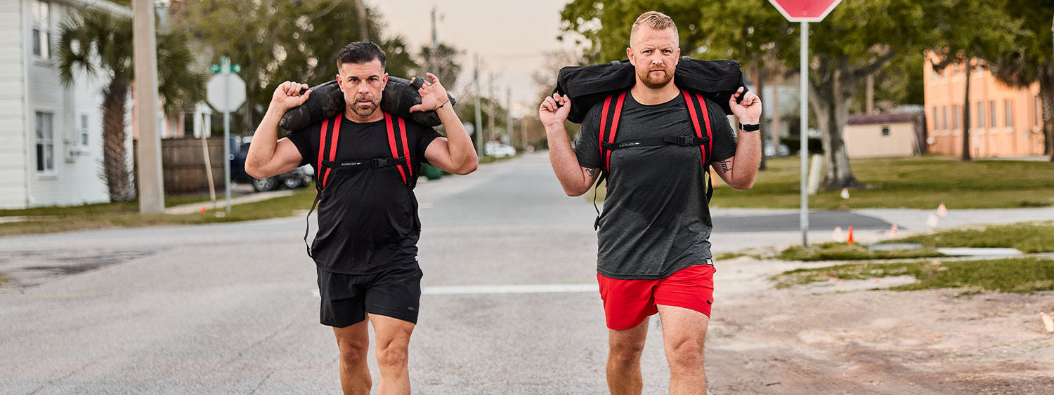 Two men walking outdoors, carrying weighted sandbags on their shoulders, wearing athletic clothing.