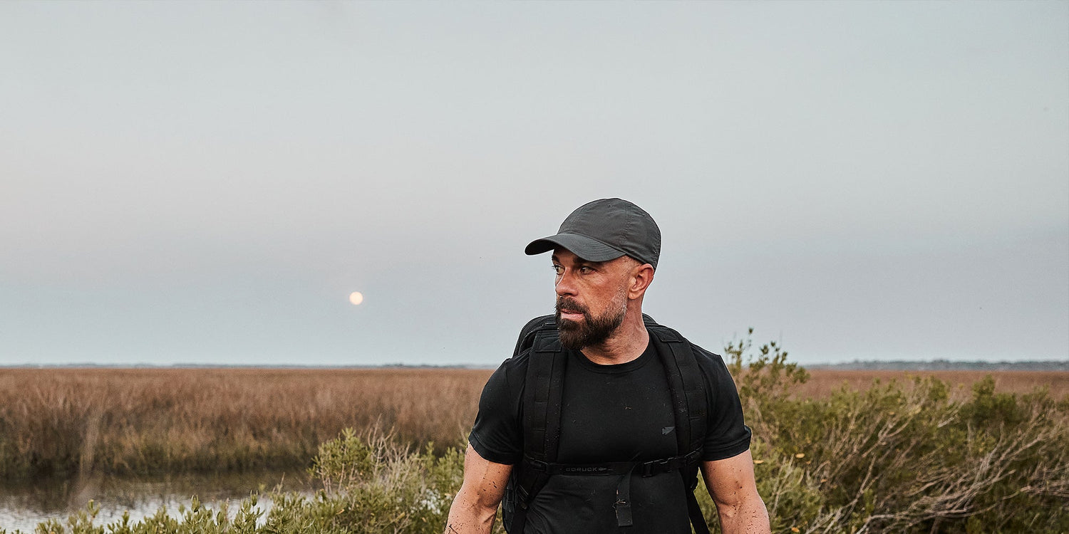 Man in black attire and cap stands in a marshland at dusk, with a backpack, under a twilight sky with a visible moon.