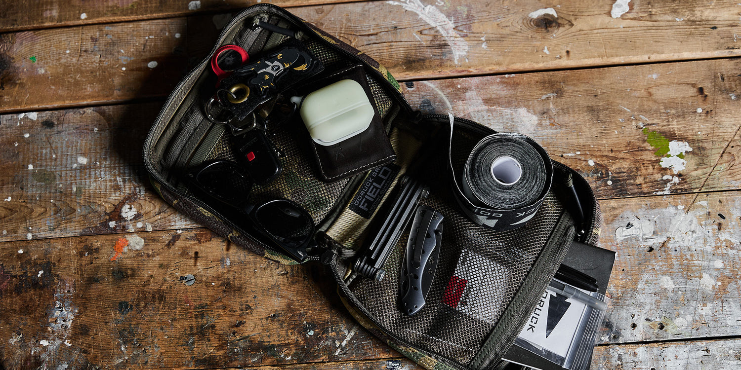 An open tactical pouch on a worn wooden table, containing tools, keys, tape, and various everyday items.