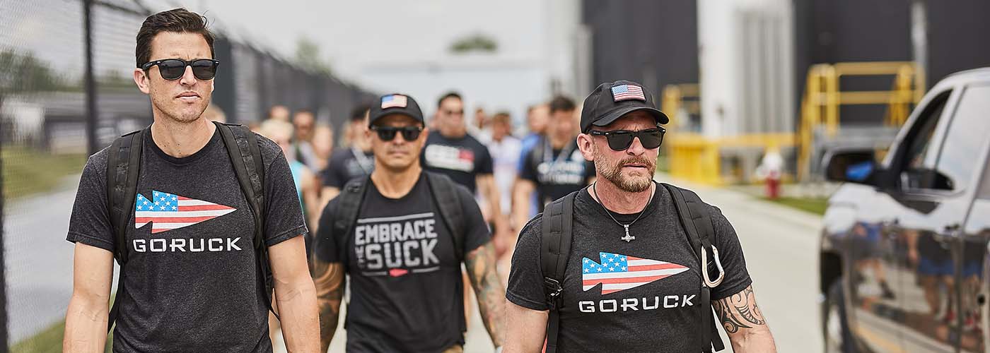 Three men in black GORUCK shirts and sunglasses walk outdoors with backpacks, others following behind them.