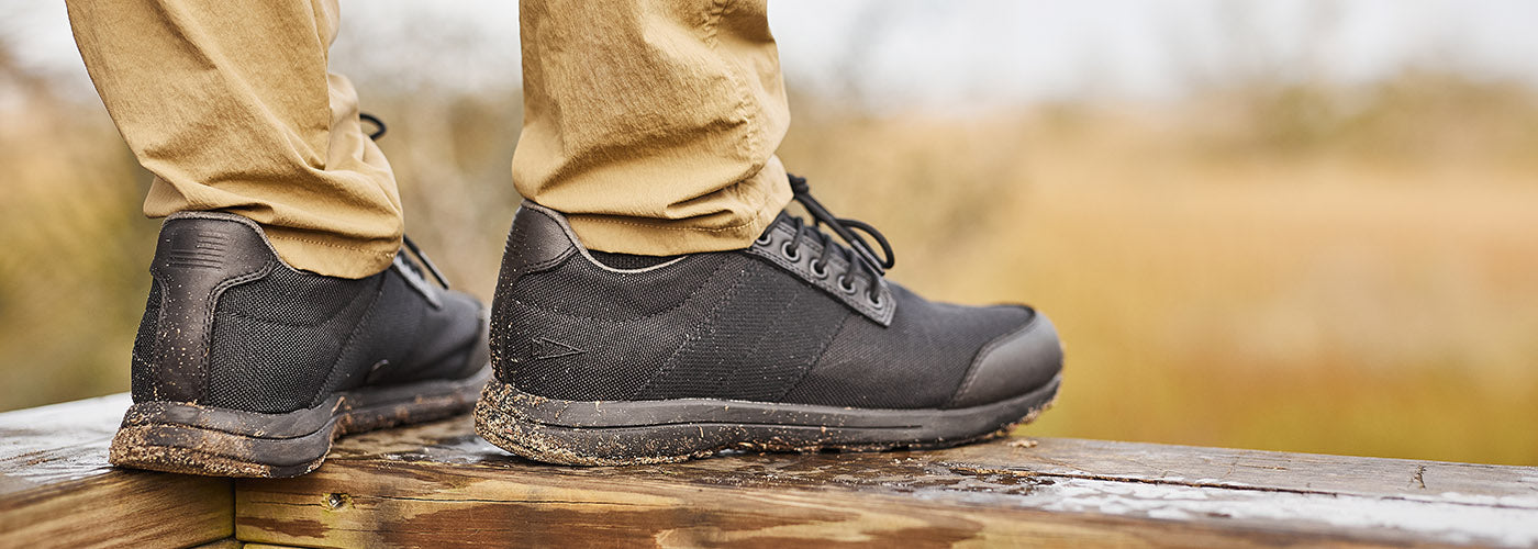 Close-up of person in beige pants wearing black shoes standing on a wooden surface outdoors.