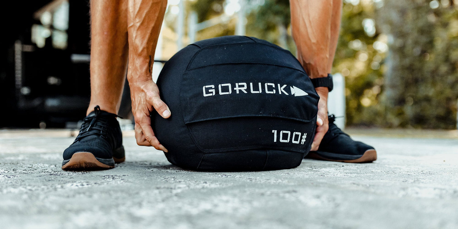 Person lifting a black GORUCK sandbag marked 100 pounds on a concrete surface outdoors.