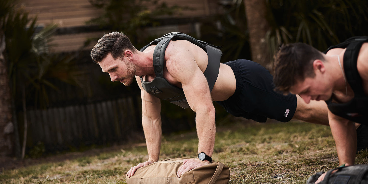Two men wearing weighted vests doing push-ups outdoors on grass near a brown sandbag.