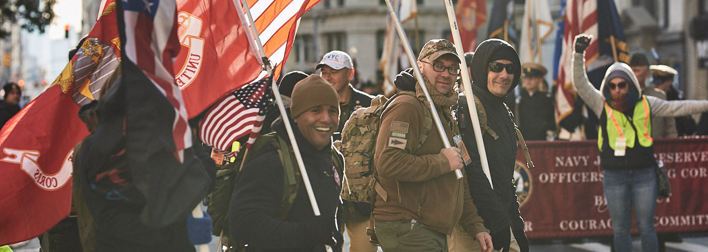 People marching with American flags, wearing casual and military-style clothing, with a large banner in the background.