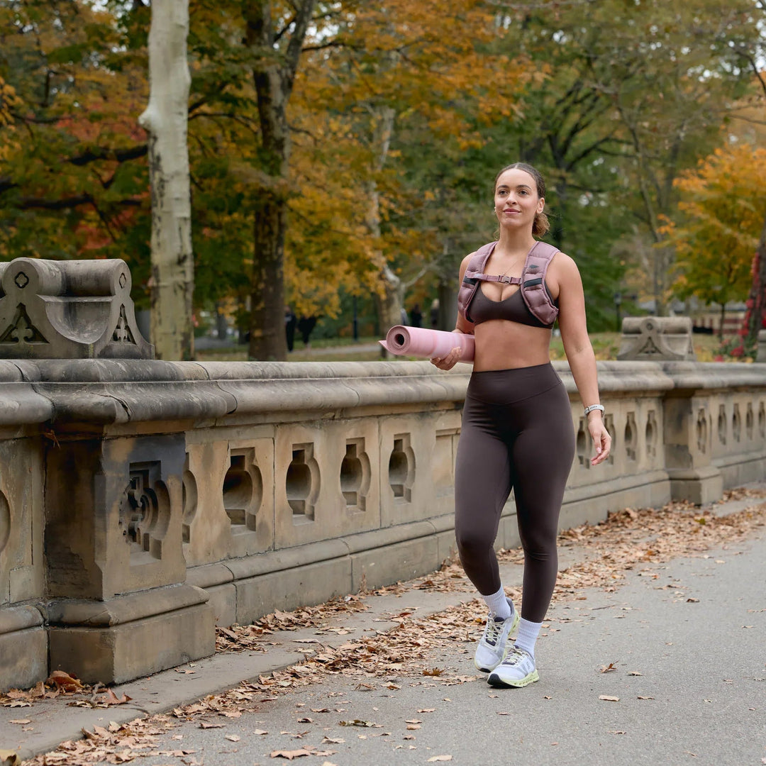 Woman rucking in GORUCK weighted vest, activewear, and holding a yoga mat outdoors in fall