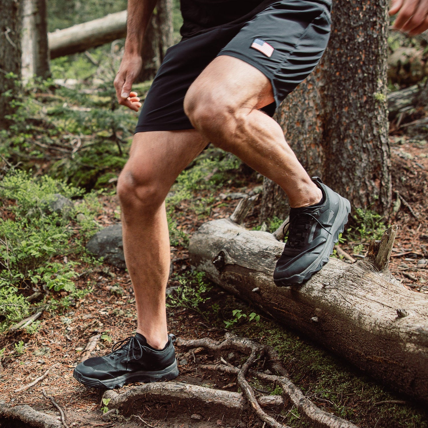 A person in black shorts and sneakers steps over a fallen log on a forest trail.