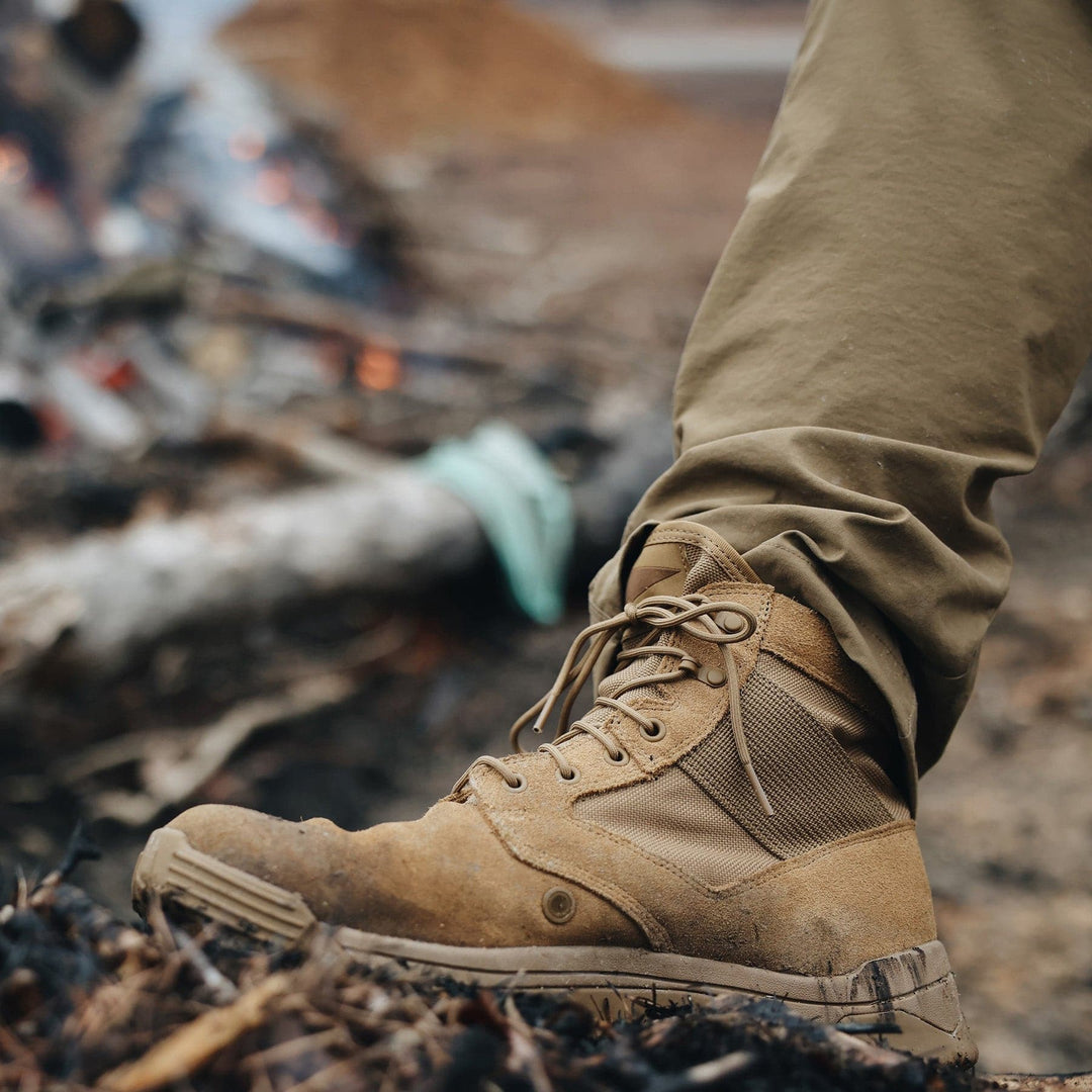 A person wearing the GORUCK MACV-2 Mid Top boots in Coyote, featuring a triple compound outsole, is standing on charred ground with olive green pants, against a blurred background of logs and flames.