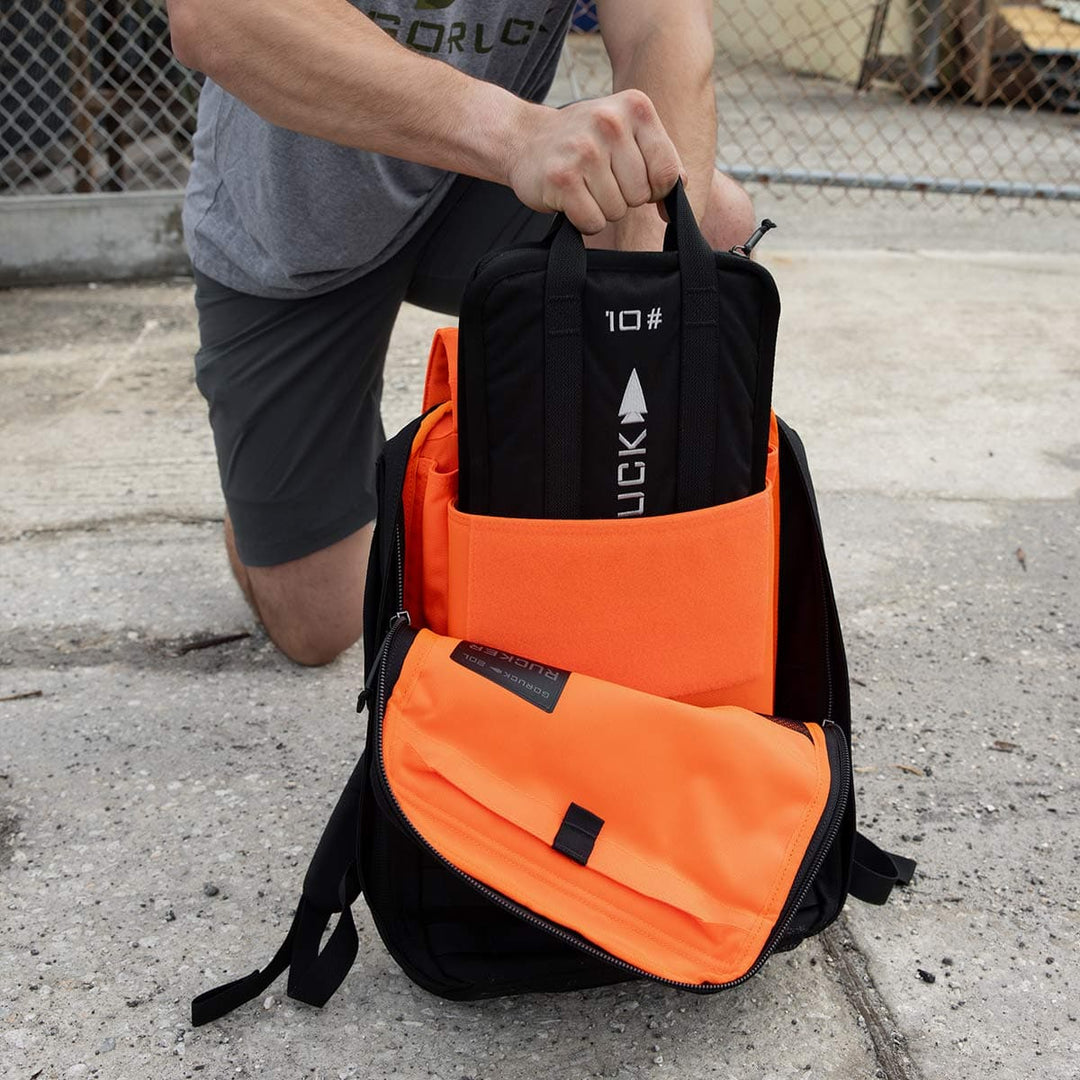 Outdoors, a person kneels carefully placing a yoga mat inside an orange and black backpack, making sure there's still space for their portable GORUCK Sand Ruck Plate.