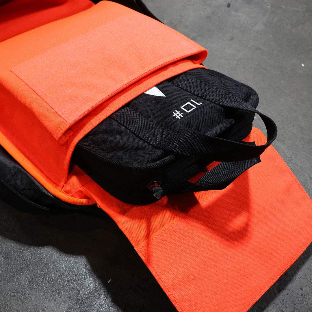 The open orange backpack on the floor reveals a black gadget case within, partially concealing a portable Sand Ruck Plate by GORUCK, perfect for rucking enthusiasts.