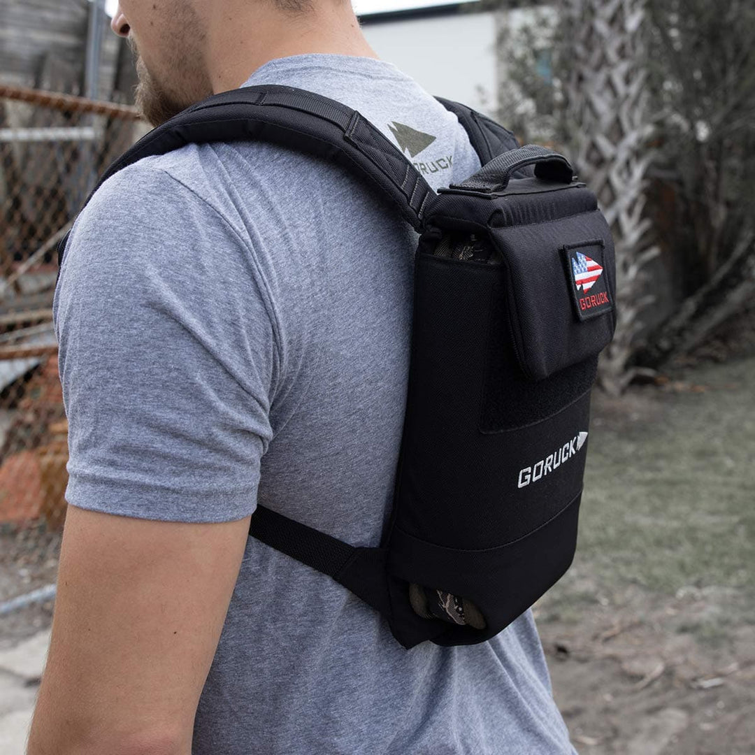 A person stands outdoors, equipped with a GORUCK backpack adorned with a patch on the back, prepared for rucking. Inside, a portable Sand Ruck Plate from GORUCK adds weight for an intensified fitness challenge.