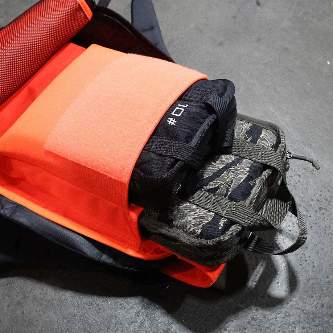 An opened orange backpack, designed for rucking, reveals its neatly packed compartments on a gray floor, with a portable GORUCK Sand Ruck Plate snugly tucked inside.