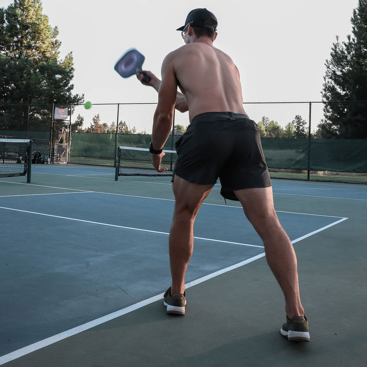 Shirtless man playing pickleball on an outdoor court, preparing to hit the ball with his paddle.